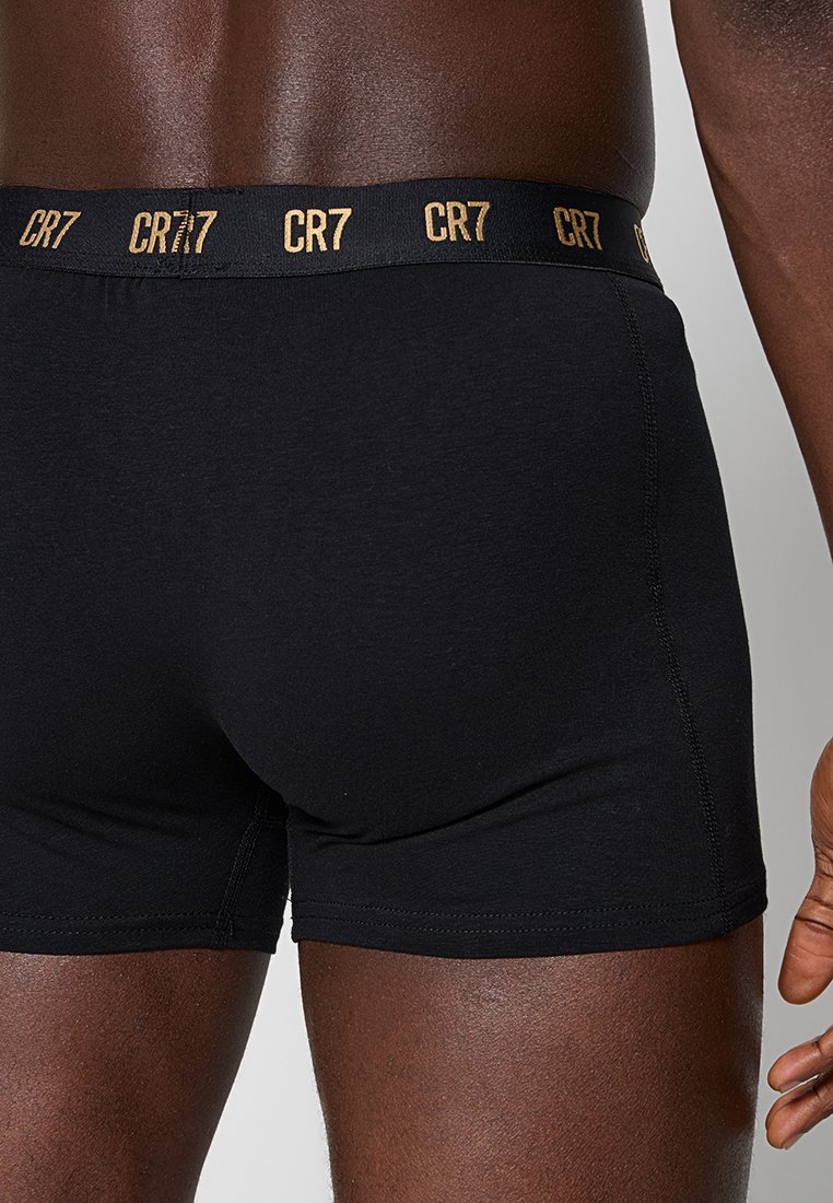 Männlicher Oberkörper und Oberschenkel, die schwarze Boxerbriefs mit einem schwarzen Bündchen tragen, das mit wiederholtem goldenen "CR7"-Branding versehen ist.