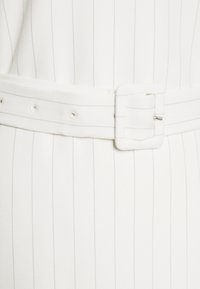 Tissu blanc avec des rayures fines verticales gris clair, doté d'une ceinture en tissu assorti avec une boucle carrée en métal et plusieurs trous ajustables.