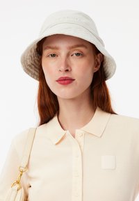 Lichtgrijze bucket hat gemaakt van zachte stof met een gestructureerde voering aan de binnenkant. Draag het met een beige overhemd met kraag en een knoopsluiting aan de voorkant.
