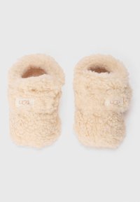 Kremfargede, fluffy tøfler laget av teksturert shearlingmateriale; rund form med et lite logomerke på siden.