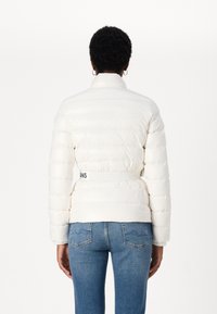 Hvit puffjakke med høy krage, horisontale sømmer og elastisk midje med logo. Kombinert med blå denimjeans.