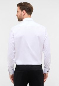 Camicia bianca a maniche lunghe con una texture morbida, colletto classico e polsini abbottonati. Abbinata a pantaloni neri su misura.