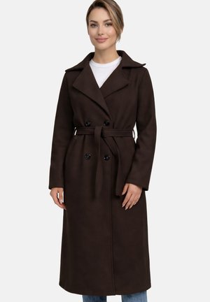 Smilende kvinde med blondt hår iført en lang mørkebrun frakke med bælte over en hvid top, stående foran en ensfarvet grå baggrund.