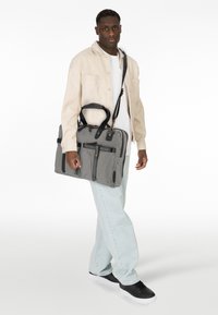 Bolsa de mensajero gris con detalles negros, múltiples bolsillos frontales y doble asa. Llevada con una chaqueta beige, camisa blanca y jeans claros.