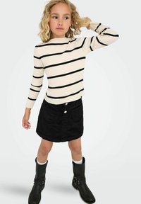 Ragazzina con capelli ricci biondi indossa un maglione crema con righe nere, una gonna nera e stivali neri, in piedi con un braccio alzato.