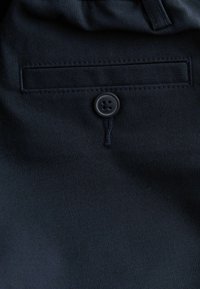 Tissu marine avec une texture lisse, présentant une poche avant et une fermeture à bouton noir unique. Les coutures sont soignées et de couleur assortie.