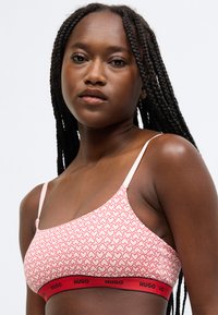 Podprsenka so srdiečkovým vzorom v červenej a ružovej farbe s nastaviteľnými ramienkami a kontrastným čiernym pásom s názvom značky „HUGO“ tučným písmom.