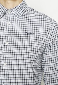 Gros plan d'une personne portant une chemise à carreaux blanche et noire boutonnée avec un col et un petit texte brodé sur la poitrine.