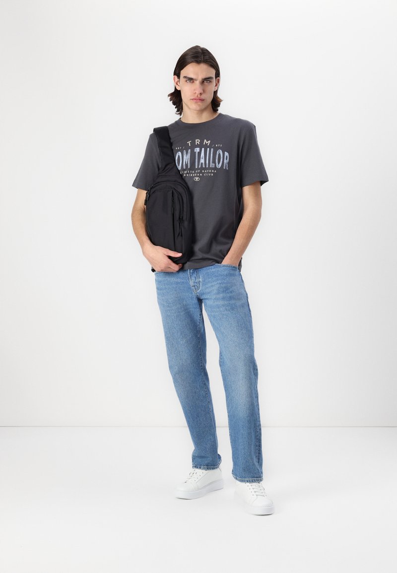 Bolso cruzado negro llevado sobre el hombro izquierdo, camiseta gris con letras azul claro, jeans azul claro y zapatillas blancas. Fondo liso.