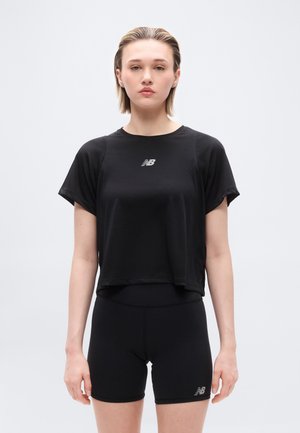 Młoda osoba ubrana w czarny sportowy krótki top New Balance i krótkie spodenki z wysokim stanem, stojąca na tle jasnej, jednolitej ściany.