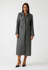 Cappotto lungo grigio strutturato con corsetto aderente, rever a sciallato, due tasche frontali e chiusura a un bottone, abbinato a tacchi alti neri.