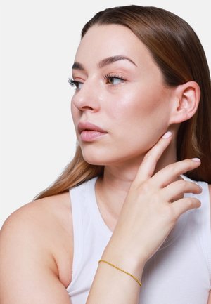 Bracciale in oro con piccoli anelli indossato su un polso, abbinato a un top senza maniche di colore chiaro. La pelle appare liscia e naturale.