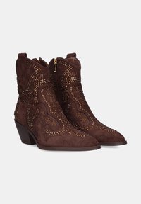 Botas de vaquero de ante marrón con punta afilada, detalles de costura y acentos de tachuelas doradas. Cuentan con un zipper lateral y un tacón bajo y apilado.