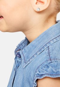 Camicia di denim con colletto, di colore azzurro chiaro e cuciture viola. Un orecchio presenta un orecchino a forma di faccina sorridente circolare.