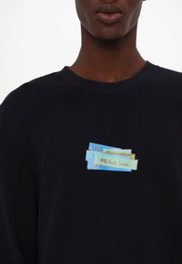 Primo piano di una persona che indossa una felpa nera con un logo colorato a strati di tessuto con la scritta "PS Paul Smith" sul petto.