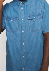 Denimová košile na knoflíky v světle modré barvě s límečkem, krátkým rukávem, dvěma náprsními kapsami a stříbrnými druky. Měkká, texturovaná látka.