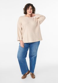 Béžový rebrovaný sveter so širokým výstrihom a dlhými rukávmi, skombinovaný s modrými džínsami s rovnými nohavicami a hnedými topánkami. Jednoduché biele pozadie.