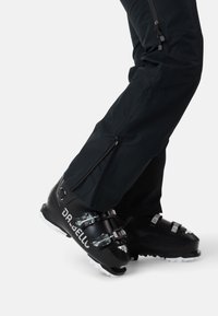 Schwarze Skistiefel mit silbernen Schnallen und strukturierter Oberfläche, kombiniert mit schwarzen technischen Hosen, die Reißverschlüsse an den Unterschenkeln aufweisen.