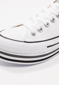 Bílé plátěné tenisky s černou gumovou podrážkou, kulatou špičkou, stříbrnými očky a černo-bílou pruhovanou podrážkou.