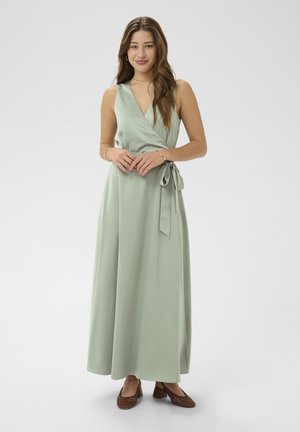 Jonge vrouw staand, gekleed in een mouwloze wikkeljurk in saliegroen met zijstrik en bruine gevlochten schoenen met blokhak, handen samengevouwen.