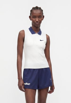 Femme aux cheveux tressés portant un polo blanc sans manches avec un col bleu marine et un short Nike bleu marine, debout devant un fond uni.