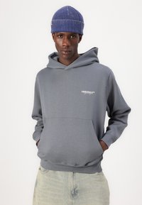 Grijze hoodie met een voorzak en ribgebreide manchetten, met witte logo-tekst op de borst. Het model draagt een blauwe ribgebreide beanie.