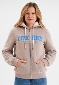 Felpa con zip di colore beige, con cappuccio foderato in pile, caratterizzata da testo blu "CREEKS", tasche frontali e polsini a costine.