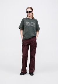 Nadrozmerné zelené tričko Nike s bielym logom, skombinované s bordovými teplákmi s bielymi bočnými pruhmi. Model má na sebe čierne slnečné okuliare a topánky.