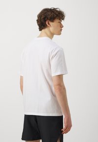 T-shirt bianca in cotone con maniche corte, colletto girocollo e design semplice. Indossata con pantaloni corti neri. Lo sfondo è semplice e di colore chiaro.
