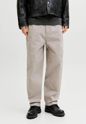 Homme portant un pantalon beige ample, des chaussures en cuir noires, une chemise noire et une veste courte en cuir noir, debout devant un fond uni.