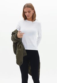 Witte langek mouw shirt met ronde hals, gecombineerd met een olijfgroen pufferjack. Zwarte leggings maken de outfit af.