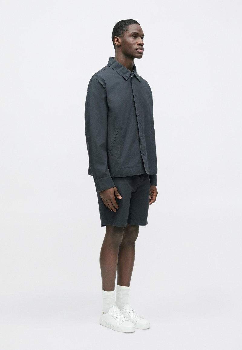 Dark gray jacket with a collar, paired with matching shorts. The model wears white sneakers and crew-length socks, standing in profile.