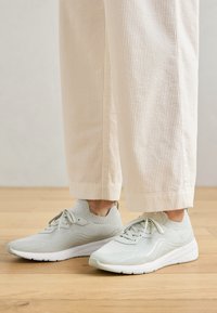 Light gray knitted sneakers with white soles worn with beige ribbed wide-leg pants on a wooden floor.