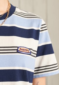 Randig t-shirt i marinblått, ljusblått och vitt. Har en cirkulär patch med "SuperDry"-logotypen. Tillverkad av bomull med en avslappnad passform.