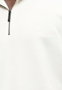 Κοντινό πλάνο ενός ατόμου που φοράει λευκό πουλόβερ με φερμουάρ και διακριτικό ανάγλυφο λογότυπο Aston Martin στο στήθος.