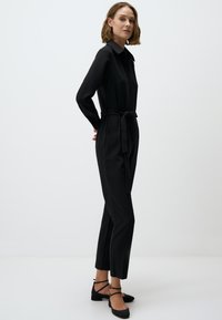 Zwarte jumpsuit met lange mouwen, een kraag, een strikceintuur en aansluitende broekspijpen. Gecombineerd met zwarte flats met enkelbandjes.