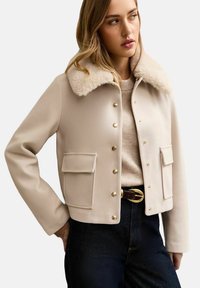 Kobieta ubrana w beżową wełnianą kurtkę z kołnierzem ze sztucznego futra, złotymi guzikami, kieszeniami z patkami, w połączeniu z jasnym swetrem i ciemnymi jeansami z paskiem.