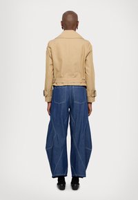 Beige cropped jasje met een brede kraag, gecombineerd met losse blauwe denim broek met contrasterende witte stiksels en een uniek paneelontwerp.