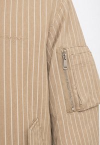 Veste beige avec fines rayures blanches verticales, dotée d'une poche zippée sur la manche supérieure et d'un emblème cousu sur la poitrine.