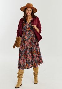 Długa sukienka w kwiatowy wzór w ciemnych odcieniach, zestawiona z bordowym kardiganem. Dodatki obejmują brązowy kapelusz oraz beżowe kozaki do kolan.