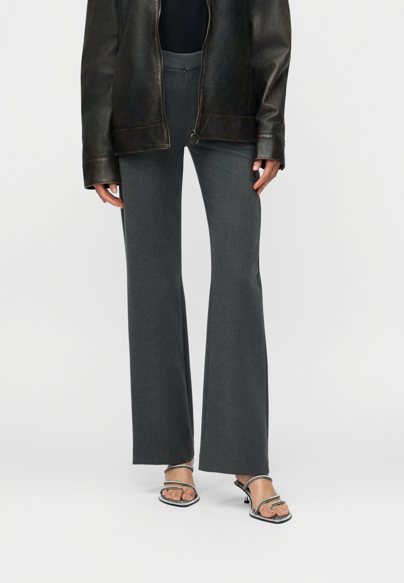 Άτομο που φοράει σκούρα γκρι παντελόνια φαρδιάς γραμμής, μαύρο τοπ, μαύρο δερμάτινο μπουφάν και λαμπερά σανδάλια με τακούνι και ανοιχτά δάχτυλα, στέκεται μπροστά σε λευκό φόντο.