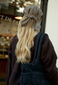 Cappello a spina di pesce marrone con un fiocco, indossato con lunghi capelli mossi. Una borsa a tracolla nera imbottita e un maglione scuro completano il look.