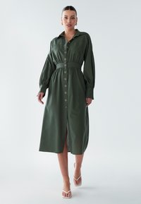 Žalia, sagomis užsegama suknelė su išryškinta juosmens linija, ilgomis rankovėmis ir vidutinio ilgio kirpimu. Yra didelės sagos ir šoninė skeltukė.