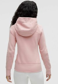 Svijetlo ružičasta dukserica napravljena od mekanog materijala, s kapuljačom na vezanje, rebrastim rukavima i uskim krojem s diskretnim logo detaljem na leđima.