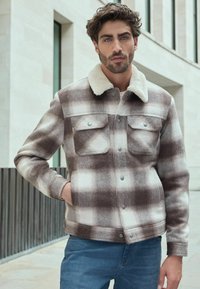 Brown and cream plaid jacket with a fuzzy collar, two chest pockets, and snap buttons, paired with blue jeans.