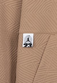 Tessuto beige con un motivo a spina di pesce testurizzato, con un'etichetta bianca che mostra un logo nero e "23" in grassetto.