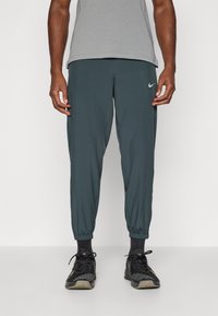 Pantaloni atletici ușori, de culoare verde închis, cu picioare îngustate și manșete elastice, având un mic logo argintiu Nike pe piciorul stâng.