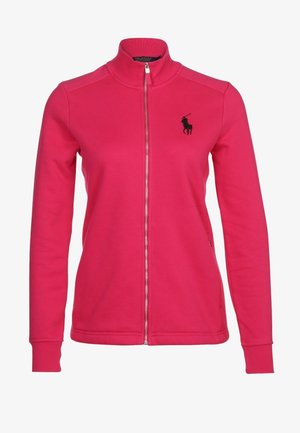 Person iført en skrigende pink jakke med lynlås, mørkeblå bukser og hvide sneakers, stående med den ene hånd i lommen mod en ensfarvet baggrund.