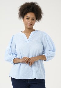 Blouse bleu clair avec un décolleté en V, des manches volumineuses et un motif à pois texturé. Tissu doux avec une coupe décontractée et des poignets froncés.