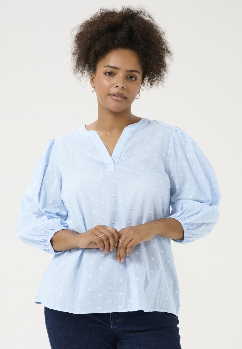 Blouse bleu clair avec un décolleté en V, des manches volumineuses et un motif à pois texturé. Tissu doux avec une coupe décontractée et des poignets froncés.